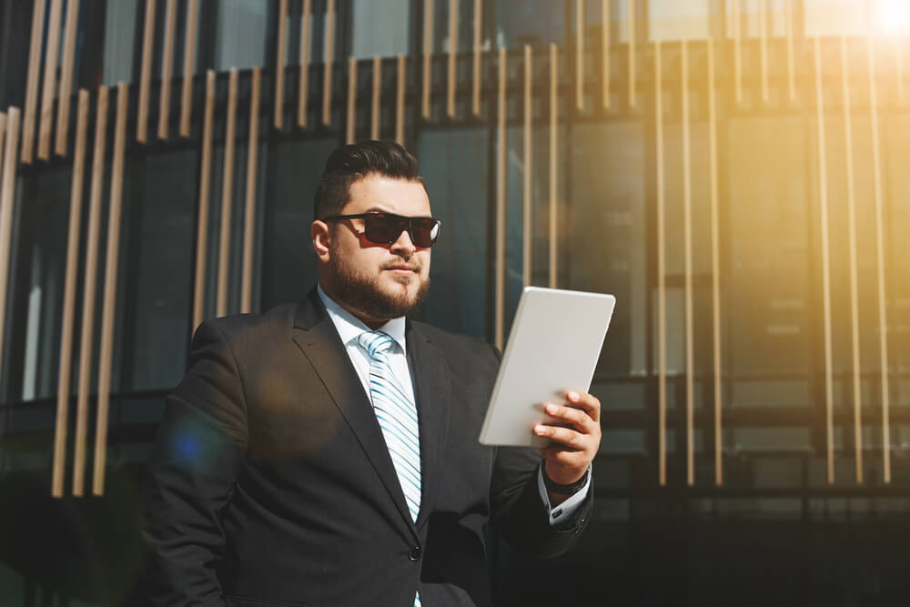 Portrait of business man reads news on tablet while standing in city financial street, adult man reading messages, stylish fat man with sunglasses using internet at sunny day outdoors, flare light
